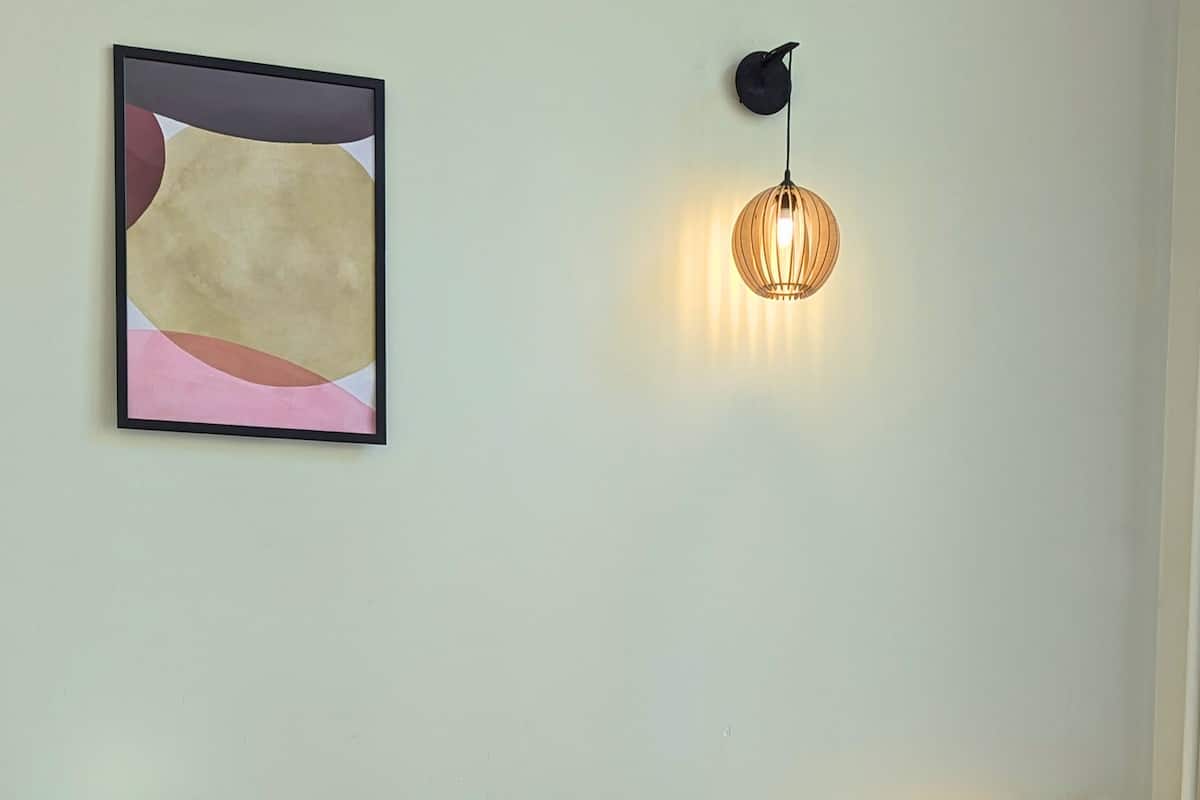 A bedroom with a bed, a desk with a chair, a wall-mounted lamp, and a framed abstract painting.