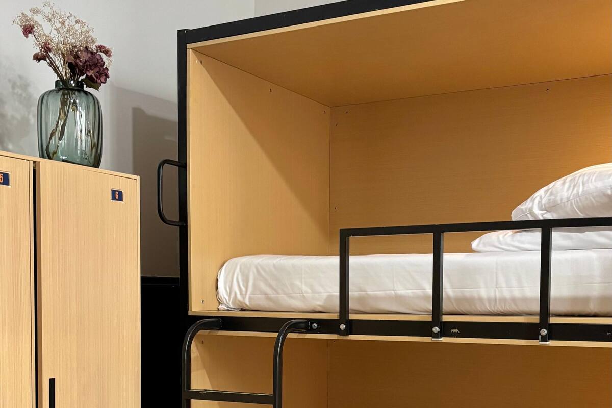 A bunk bed with a wooden frame and a single mattress. There is a small cabinet with two drawers and a vase with flowers on top.