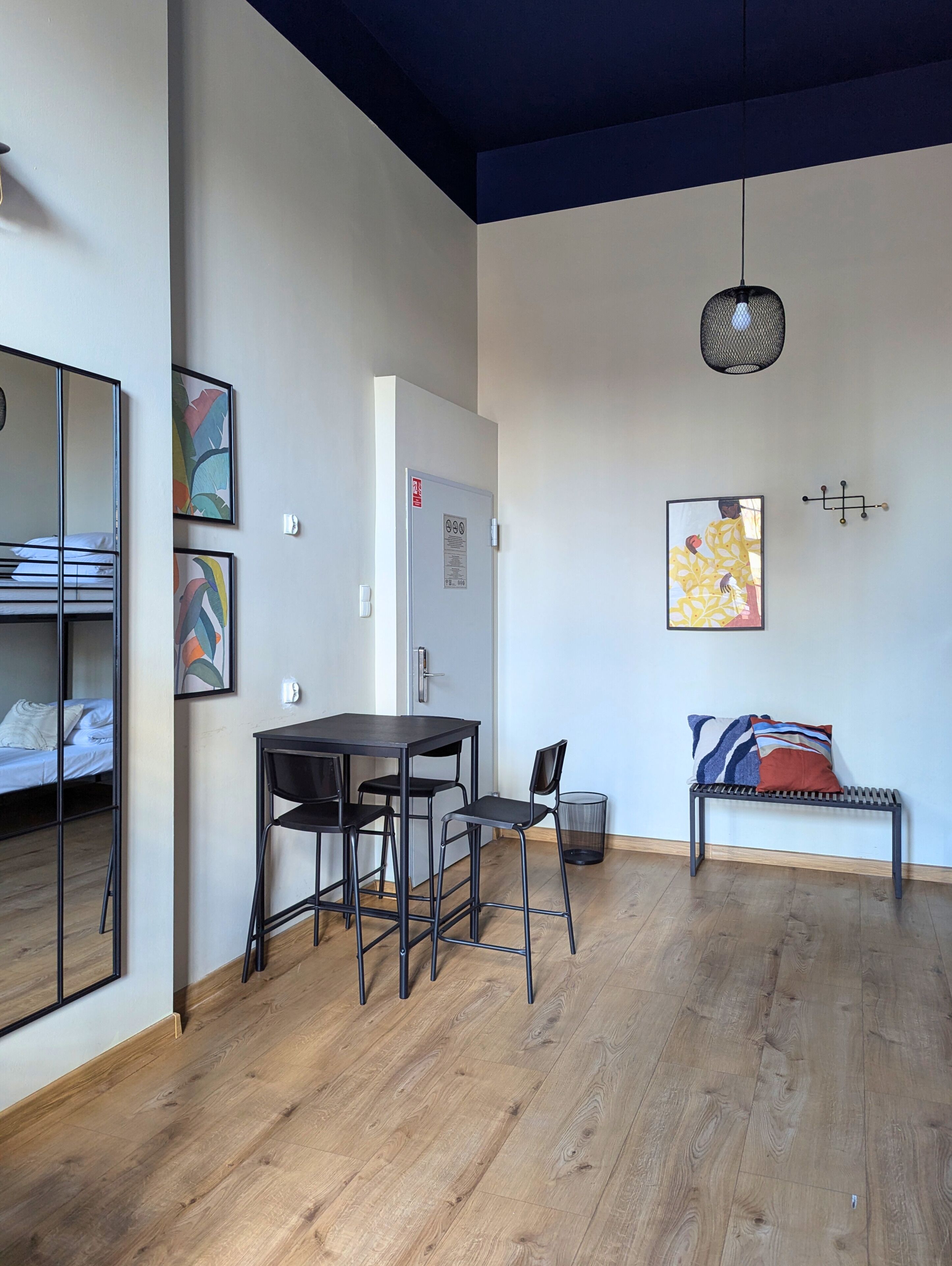 A room with a wooden floor, a dining table with chairs, a bench with cushions, a hanging light fixture, and artwork on the walls.