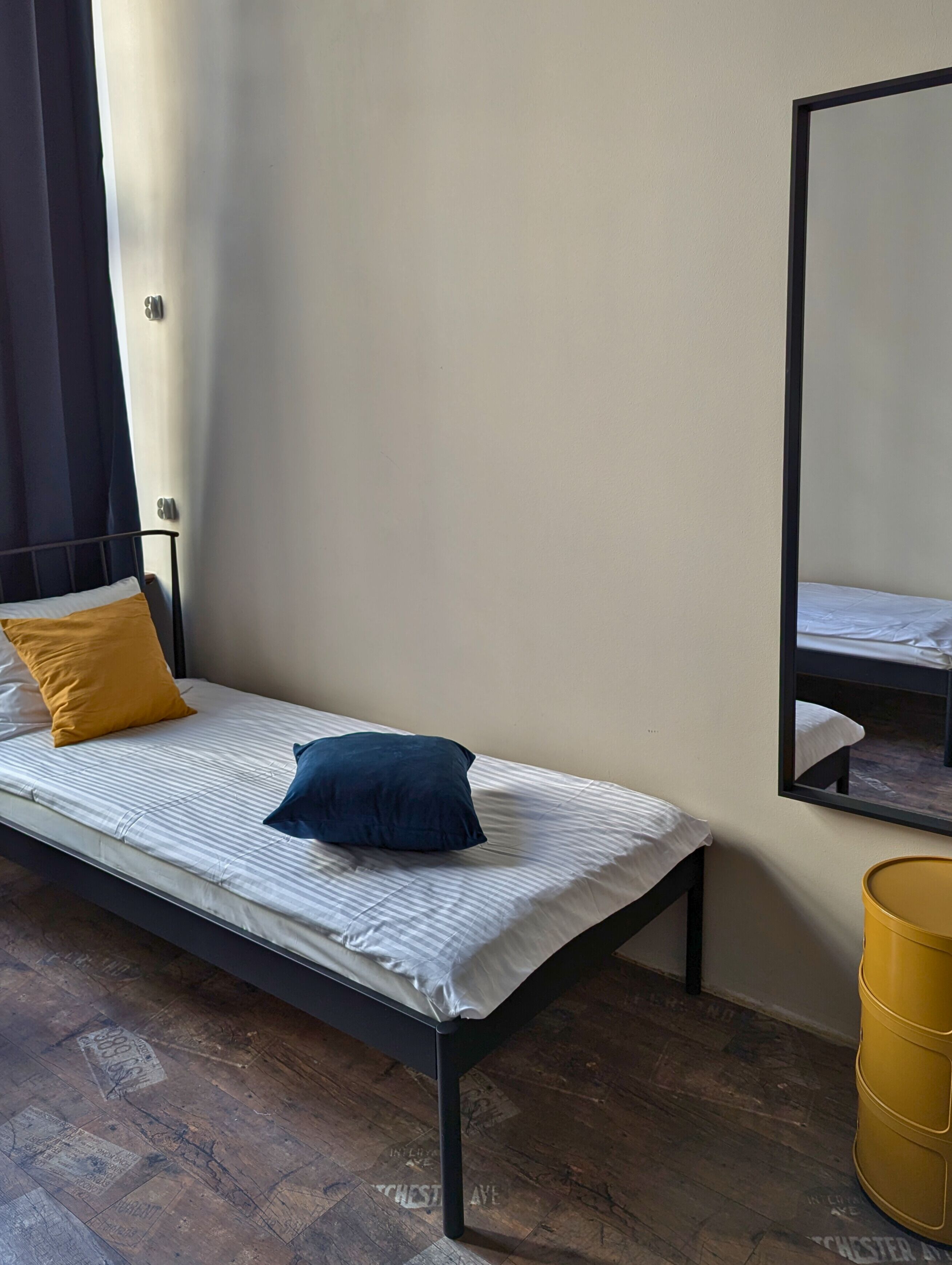 A single bed with a striped mattress, a blue pillow, and a mustard-colored pillow. A large mirror is on the wall, and a yellow trash bin is visible.