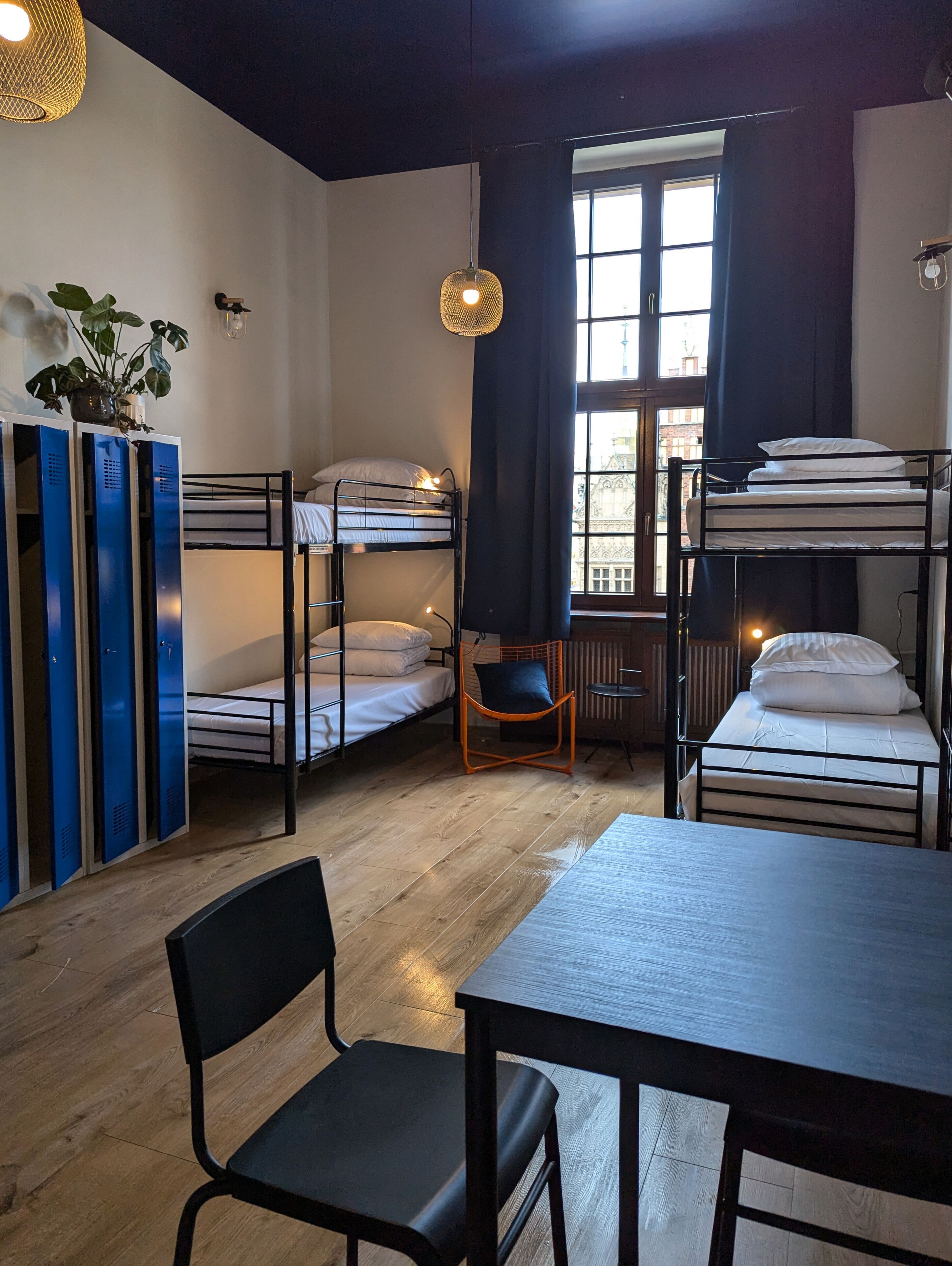 A dormitory room with bunk beds, a small table, a chair, a radiator, and a window with curtains.