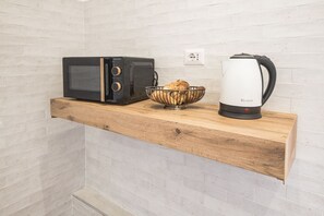 A kitchen with a microwave, a bowl of bread, and a kettle on a wooden shelf.