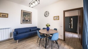 Une pièce avec un canapé bleu, une table ronde en bois entourée de chaises, une horloge et des tableaux encadrés accrochés aux murs.