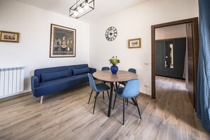 A room with a blue sofa, a round wooden table with chairs, a clock, and framed pictures on the walls.