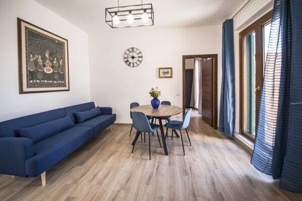 A modern living room with a blue sofa, a wooden dining table with chairs, a clock, and a framed picture on the wall.