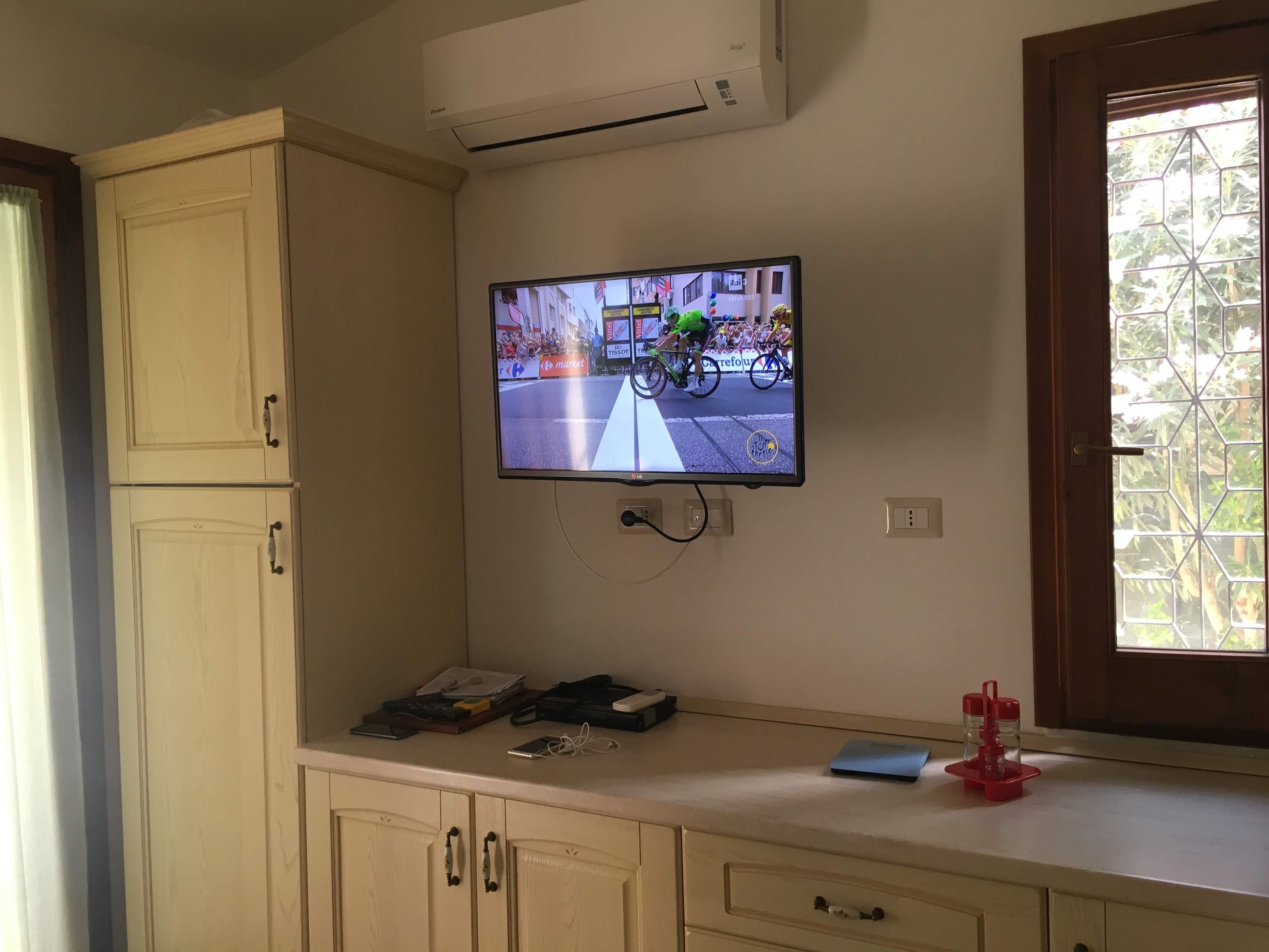 Ein Zimmer mit einem an der Wand montierten Fernseher, der eine Radveranstaltung zeigt, einem Fenster mit Blick auf Grünflächen und einer Küchenzeile mit Schränken und einer Arbeitsfläche.