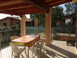 Ein überdachter Patio-Bereich mit Tisch und Stühlen, ein grüner Zaun und ein Gebäude im Hintergrund.