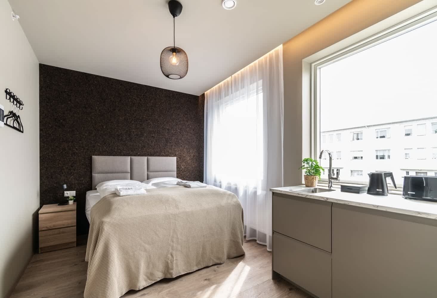 A modern bedroom with a large bed, a small bedside table, a hanging light fixture, a window with sheer curtains, and a view of buildings outside.