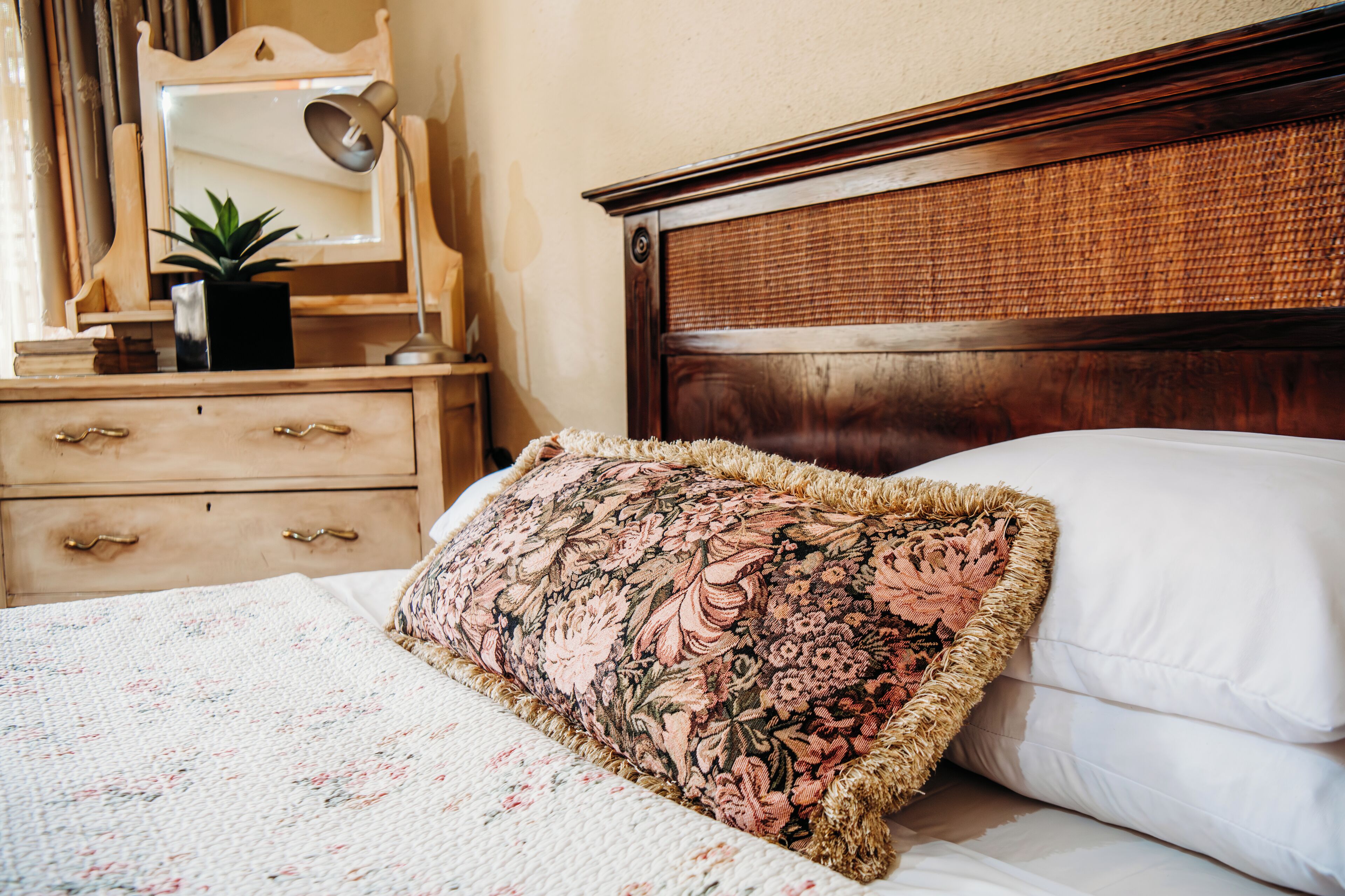 Una cama con un cojín floral, una mesita de noche con una lámpara y un espejo.