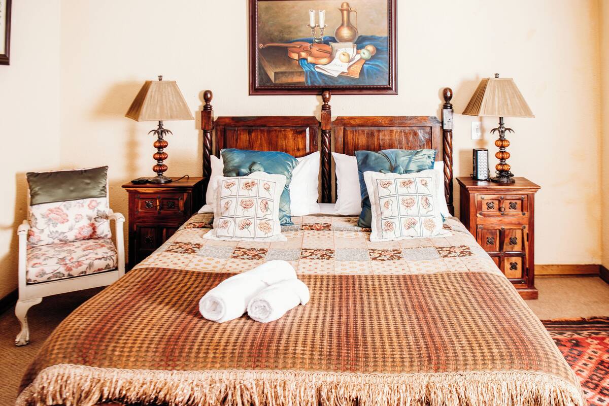 Ein ordentlich gemachtes Bett mit karierter Tagesdecke, zwei Kissen mit Blumenmuster und zwei aufgerollte weiße Handtücher. Auf jeder Seite des Bettes steht ein Nachttisch mit einer Lampe, und links vom Bett ein Sessel mit Blumenkissen.
