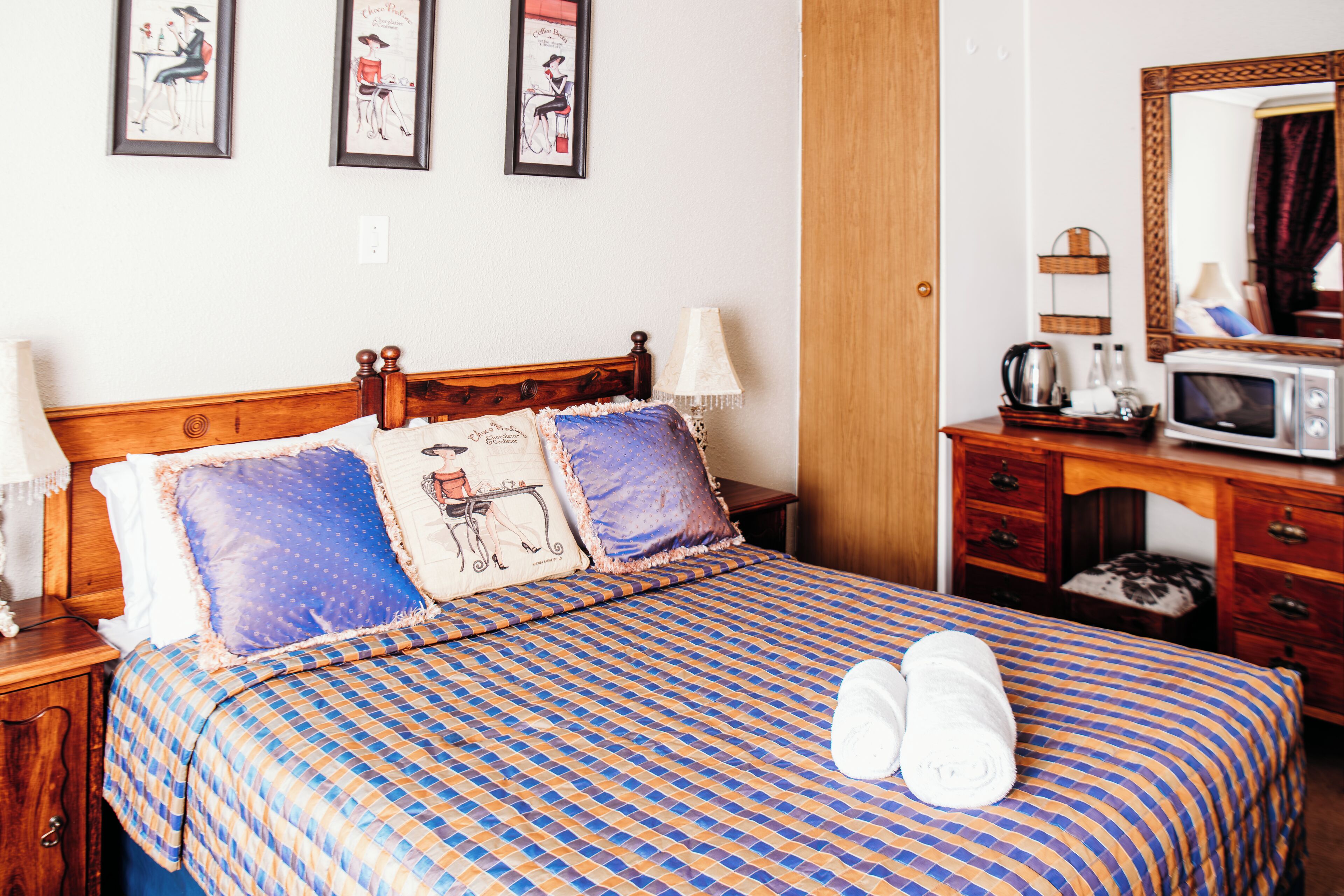 A hotel room with a checkered bedspread, a wooden headboard, two pillows, a bedside table with a lamp, a dresser with a mirror, and a microwave.