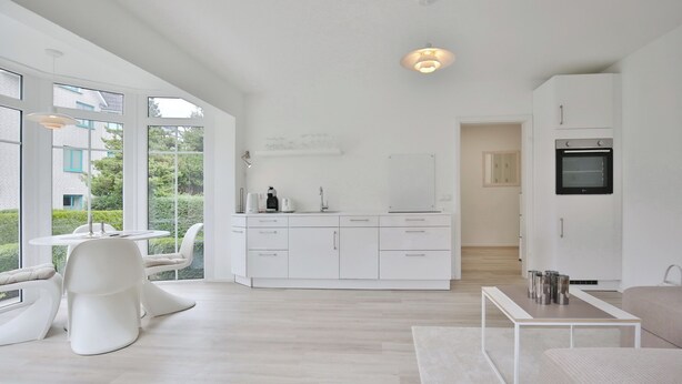 Une cuisine moderne avec des meubles blancs, un coin repas doté d’une table ronde et de chaises, et une vue sur l’extérieur grâce à de grandes fenêtres.