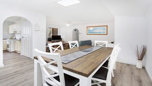 A dining area with a wooden table and white chairs, a kitchen in the background, and a painting on the wall. - My Coastal Getaway - Kiama (Kiama)