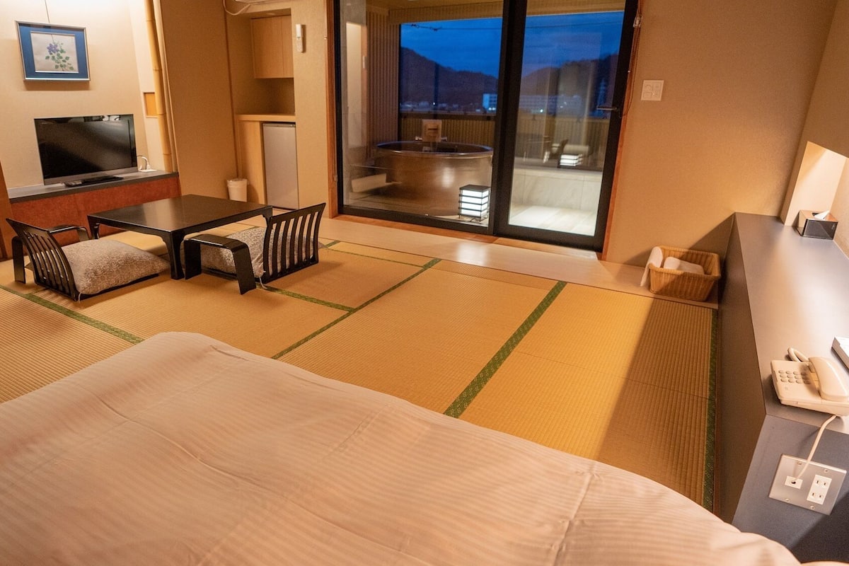 A hotel room with tatami flooring, a bed, a desk with a phone, a TV, and a sliding glass door leading to a balcony.