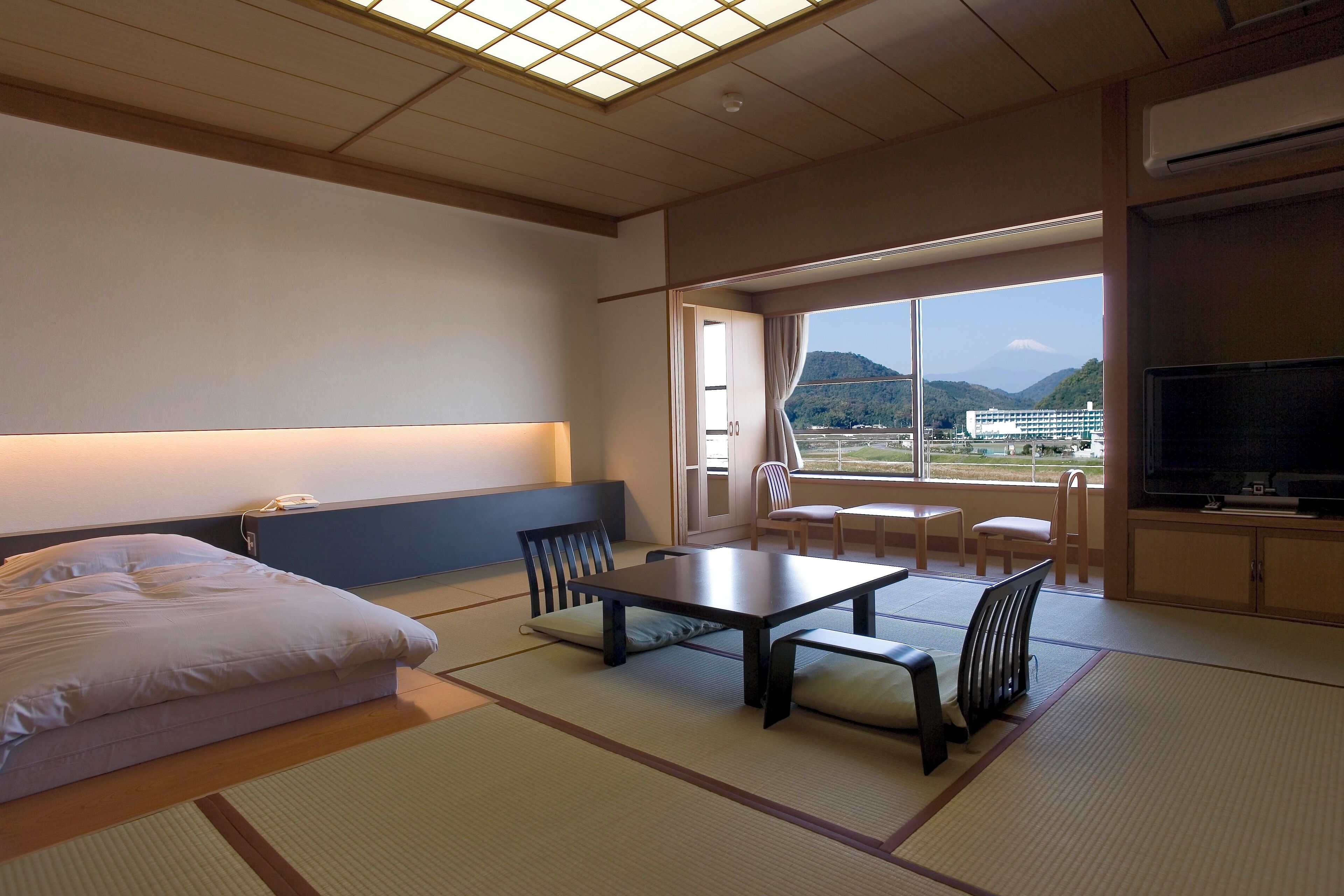 Une chambre d’hôtel avec des tatamis, un lit, une table avec des chaises, une télévision et une vue sur les montagnes à travers de grandes fenêtres.