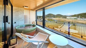 A modern balcony with a view of a river and hills, a seating area with a fire pit, and a glass door leading to a room.