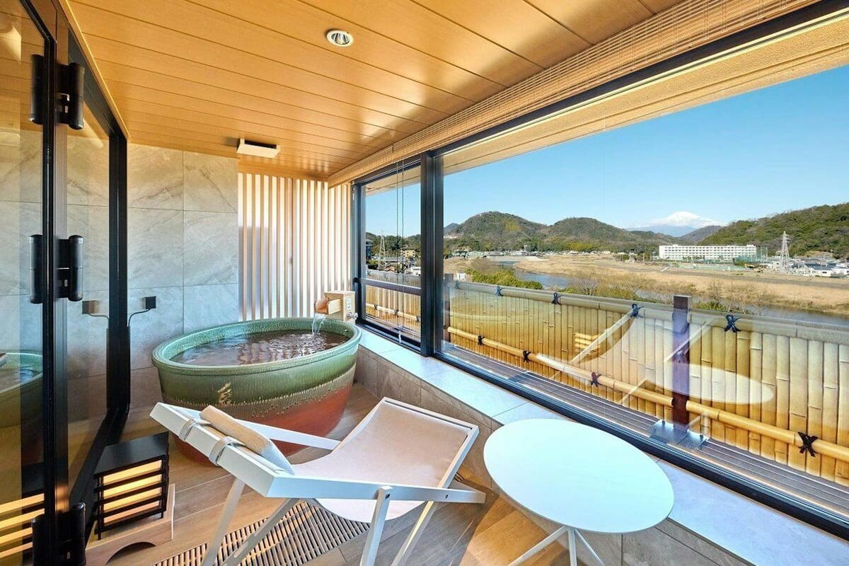 Een modern balkon met uitzicht op een rivier en heuvels, een zithoek met vuurkorf, en een glazen deur die naar een kamer leidt.