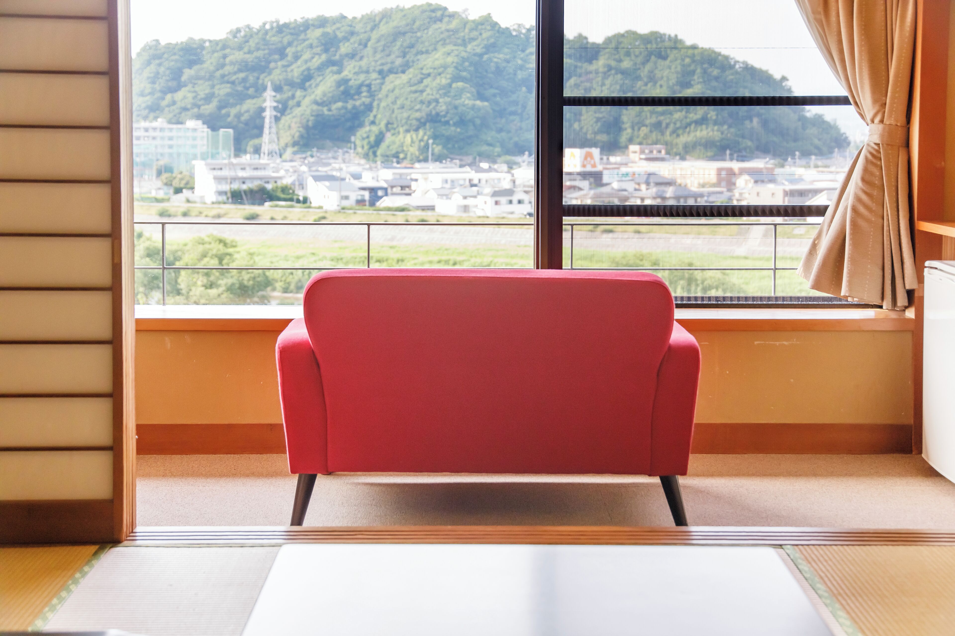 Una habitación con una silla roja, un ventanal con vistas a una ciudad y colinas, y suelo de esteras de tatami.