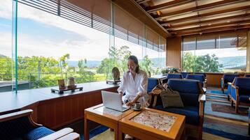 A spacious lounge area with a large window offering a view of greenery and hills. There is a wooden table with a laptop and a cup, and blue upholstered chairs.