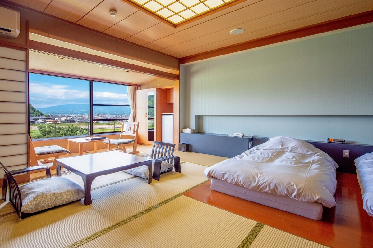 A hotel room with tatami flooring, a bed, a desk, and a view of the city.