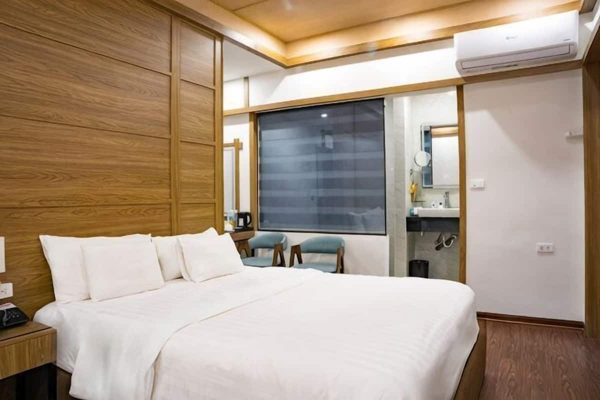 A hotel room with a large bed, a wooden headboard, a window with blinds, a small table with a chair, and a bathroom visible through a glass door.