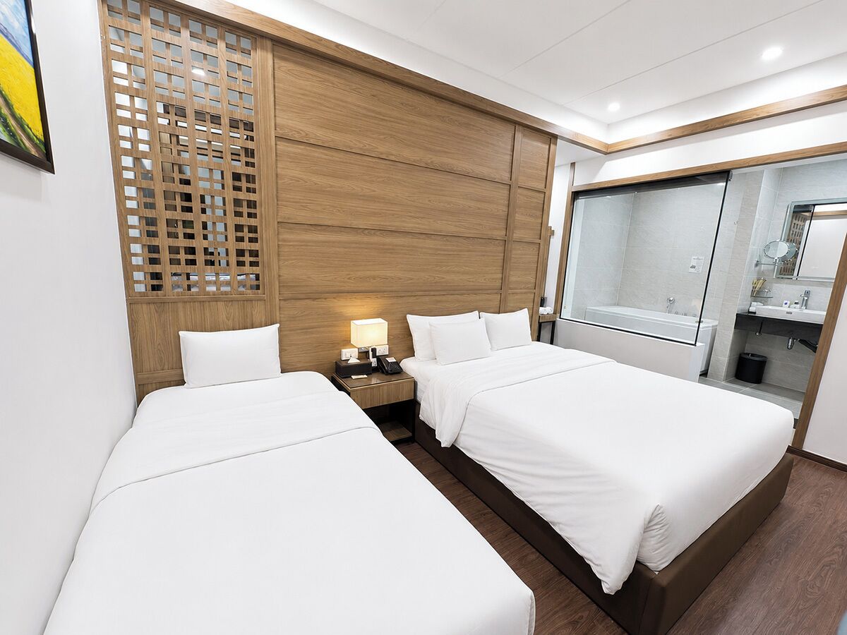 A hotel room with two beds, a wooden headboard, a nightstand with a lamp, and a bathroom with a mirror and sink.