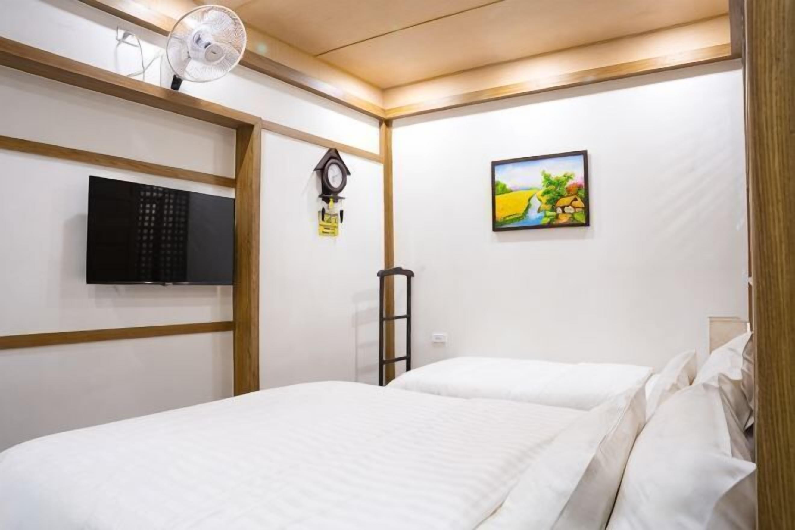Habitación de hotel con cama, televisión, reloj, un cuadro en la pared y ventilador.