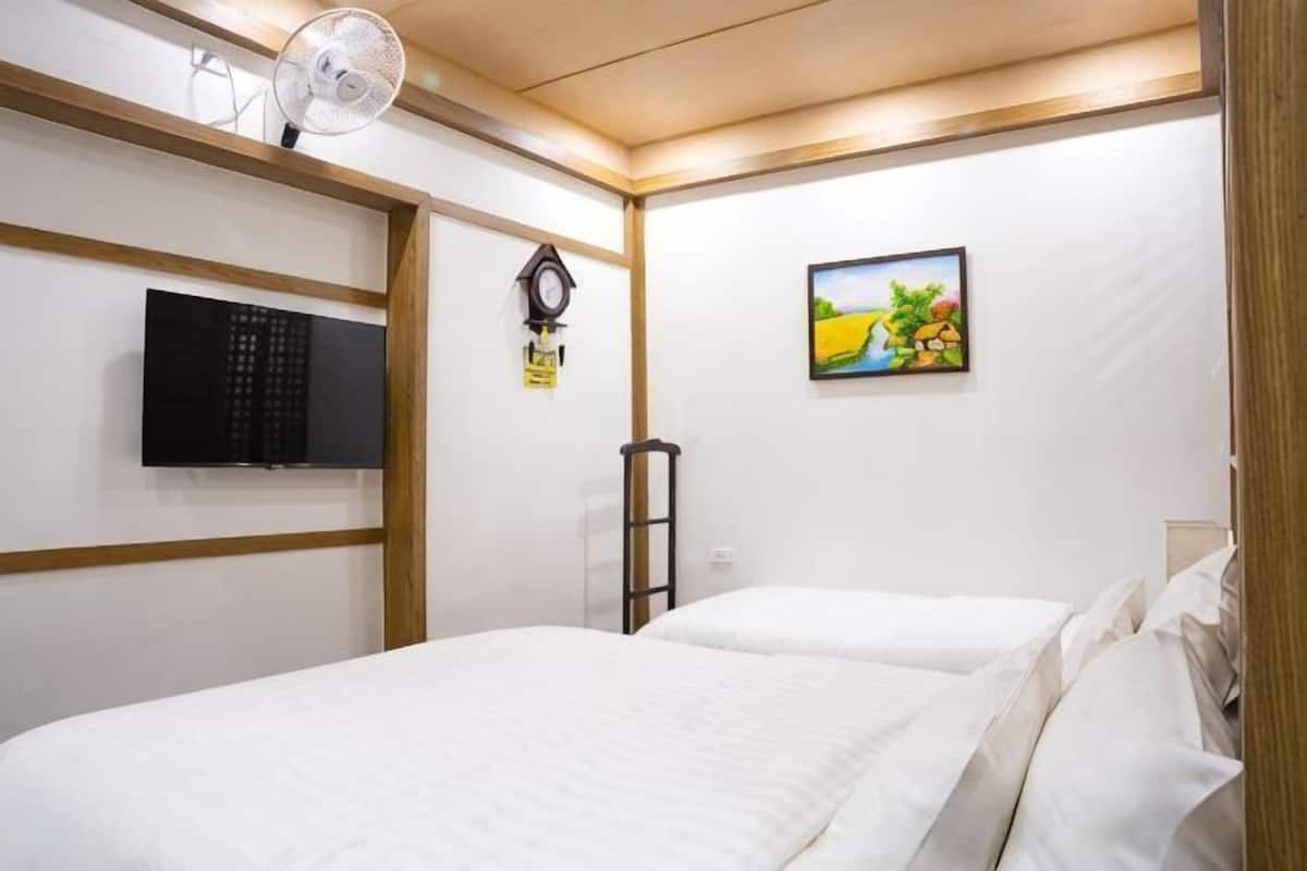 Ein Hotelzimmer mit Bett, Fernseher, einer Uhr, einem Bild an der Wand und einem Ventilator.