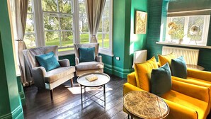 Una habitación con dos sillones, un sofá amarillo, una mesita redonda y vistas a un jardín a través de amplios ventanales.
