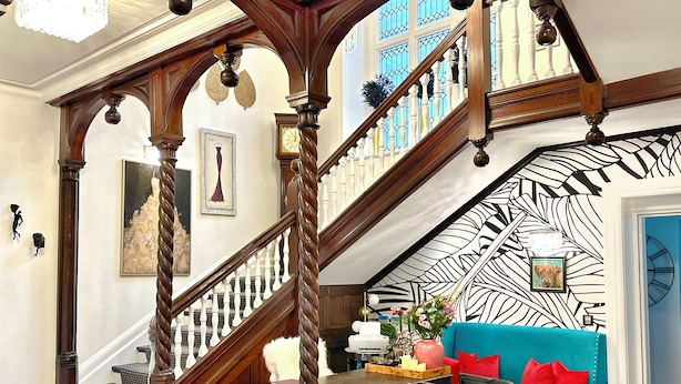 Una imponente escalera con balaustres de madera y un candelabro, que conduce a una habitación con un sofá azul, una mesa de centro y un papel tapiz estampado.