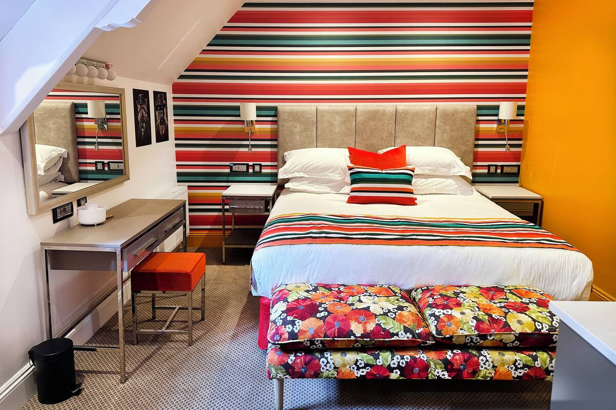 A hotel room with a striped wall design, a bed with floral bedding, a desk with a chair, and a small stool.