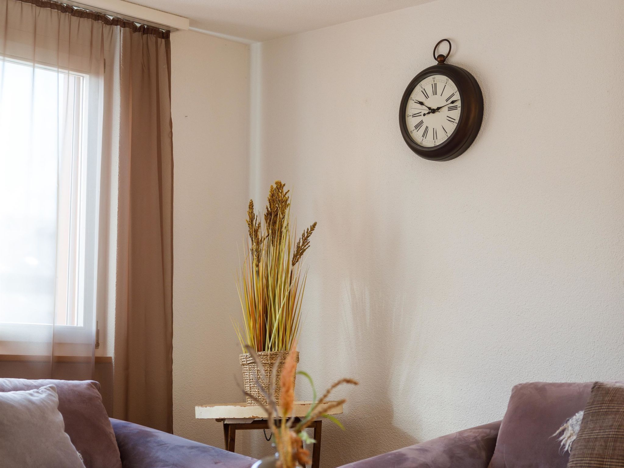 Una sala de estar con un reloj de pared, una ventana con cortinas y una mesita con una planta.