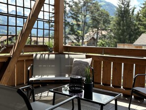 Eine Holzterrasse mit einem Glastisch, einer Bank und einem Stuhl. Auf dem Tisch stehen Blumentöpfe.