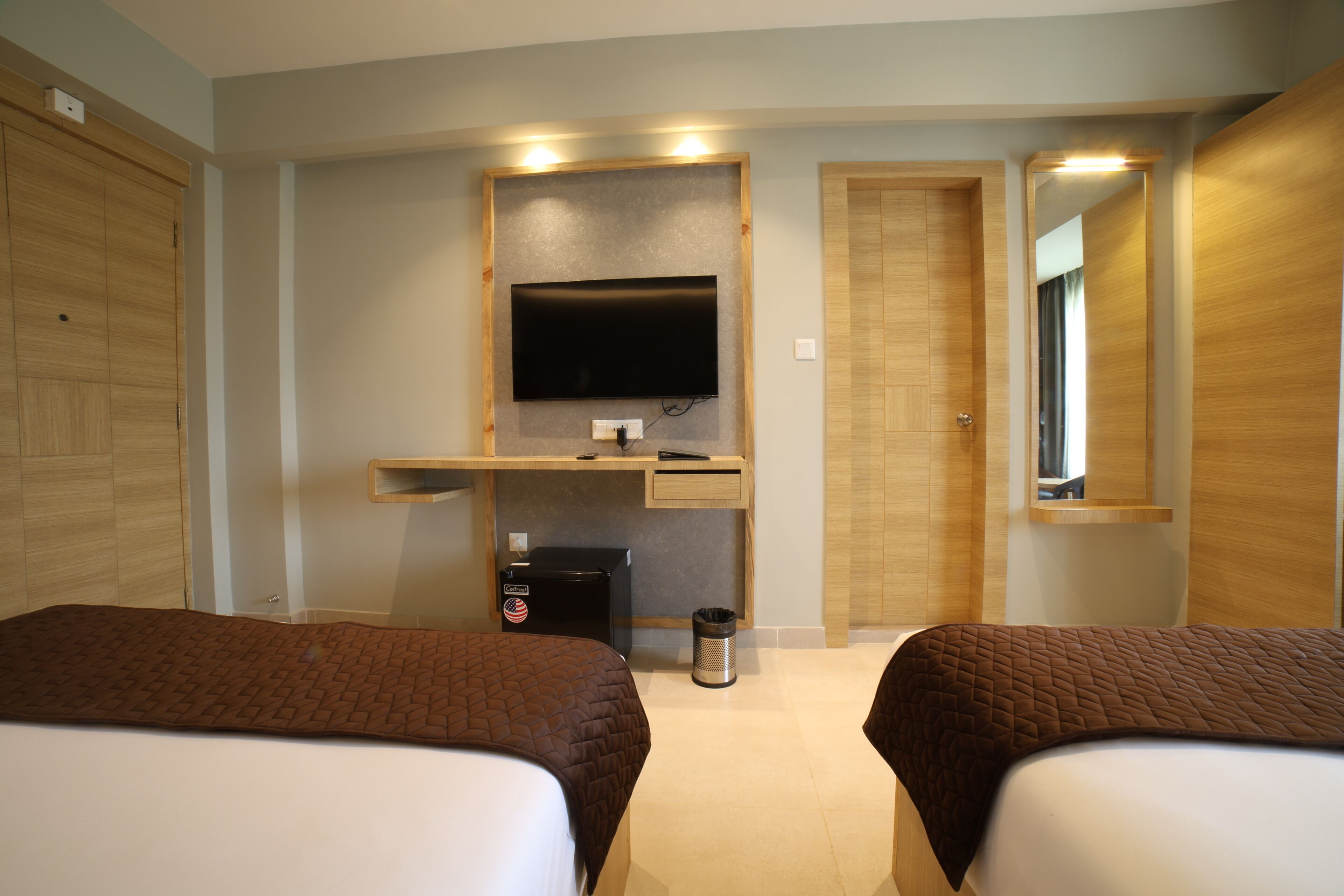 Habitación de hotel con dos camas, un televisor montado en un panel de madera, un escritorio con computadora y un bote de basura.