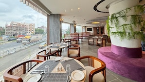 A restaurant with tables set for dining, large windows offering a view of the city, and a decorative pillar with greenery. - Northin Hotel Kolkata Airport (Barakpur)