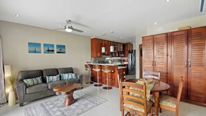 Ein modernes Wohnzimmer mit einer grauen Couch, einem Holztisch und einem Essbereich mit Holztisch und Stühlen. Es gibt eine Küche mit Holzschränken und einem Kühlschrank.