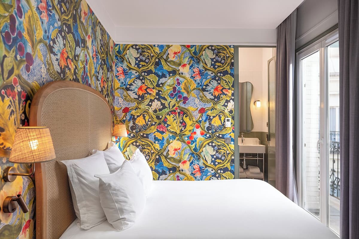 A hotel room with a bed, a headboard, a lamp, and a colorful floral wallpaper.