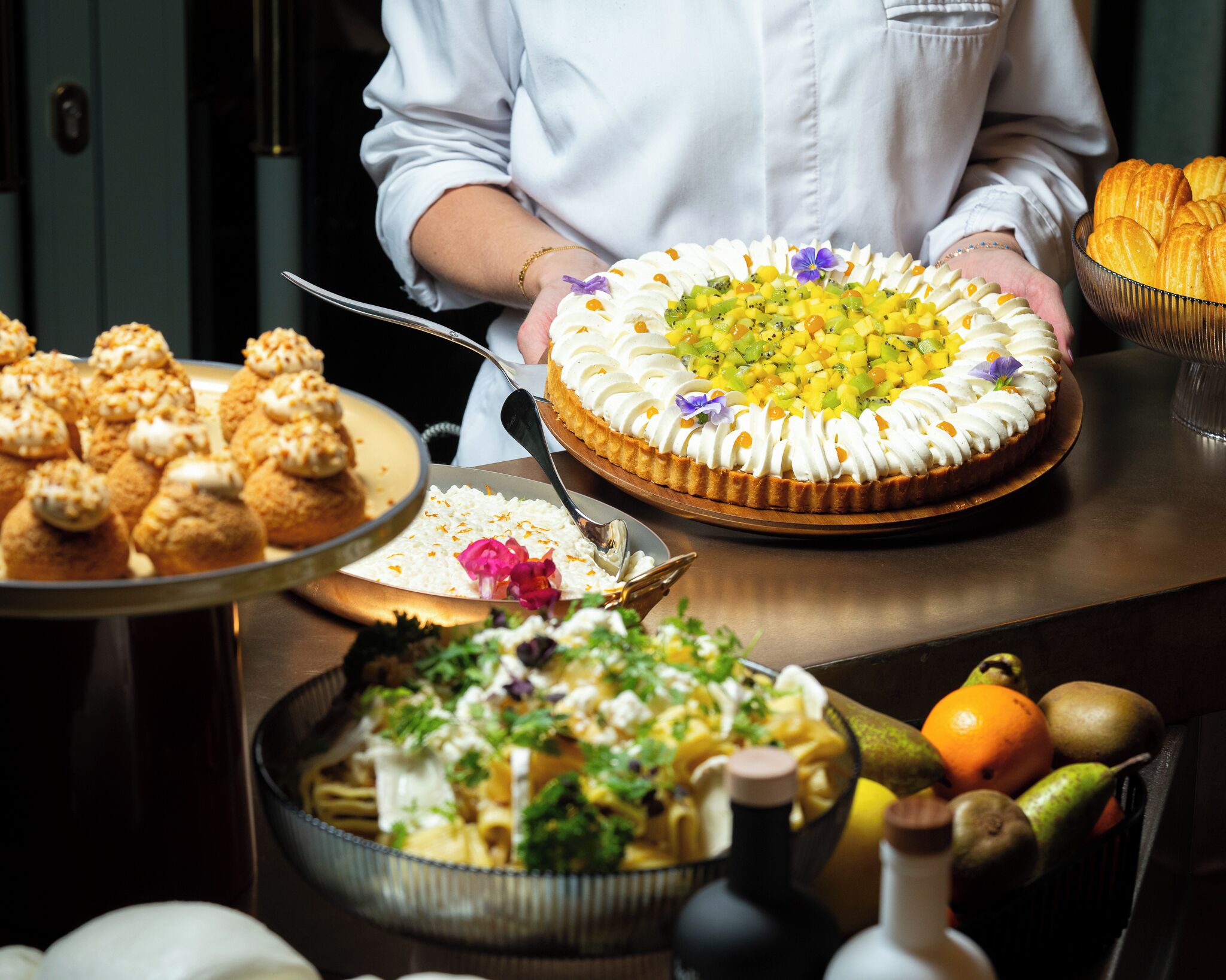 A chef holding a large fruit-topped cake, with other desserts and a fruit bowl on a table.