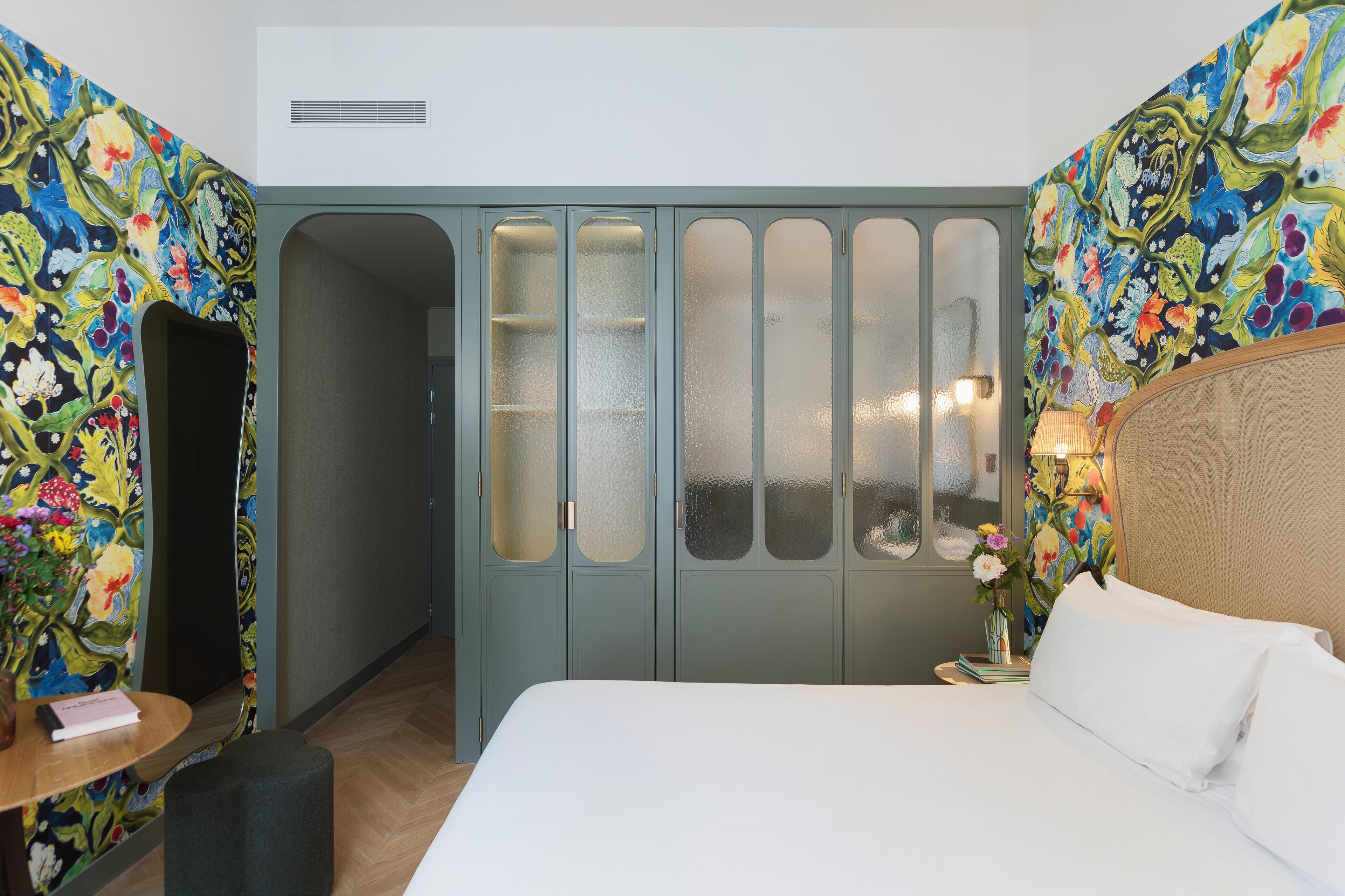 Una habitación de hotel con cama, tocador con espejo, una pequeña mesa con un libro y pared cubierta de papel tapiz floral.