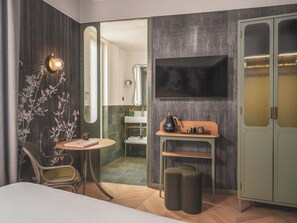 A modern hotel room with a wooden floor, a round table with a book, a chair, a TV mounted on the wall, and a wardrobe with frosted glass doors.