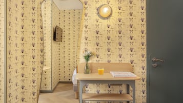 Una habitación con papel tapiz estampado, un escritorio de madera con un jarrón y un vaso de zumo de naranja, un espejo y una puerta.
