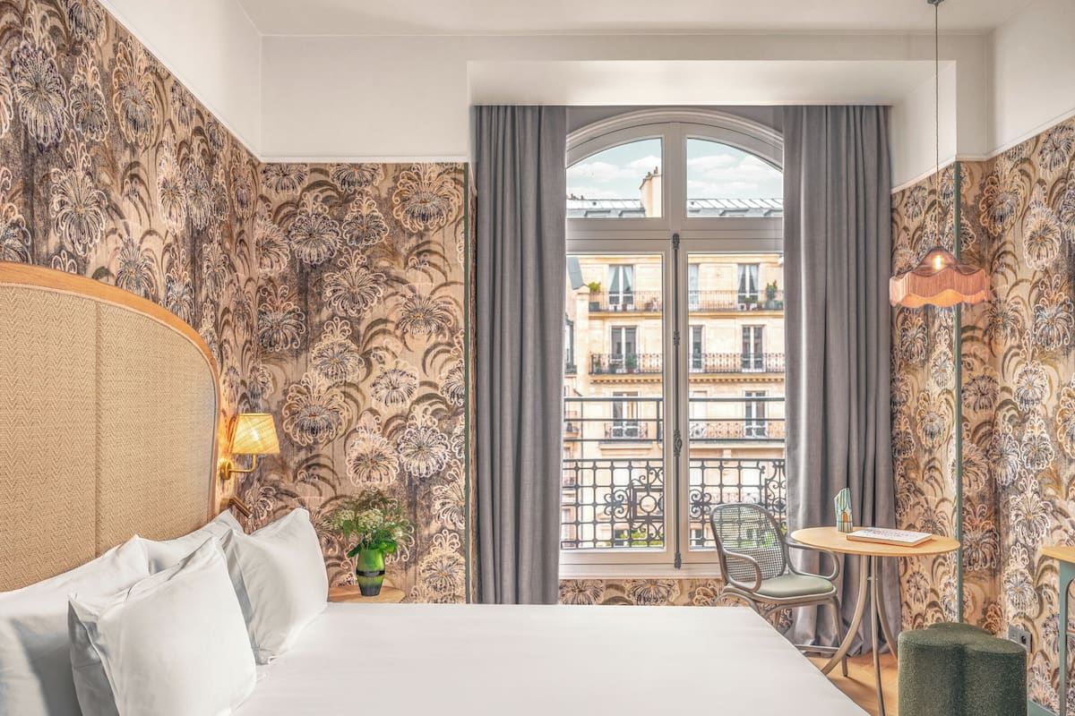 A hotel room with a large bed, a small table with a chair, a potted plant, and a view of the city through a window with a balcony.