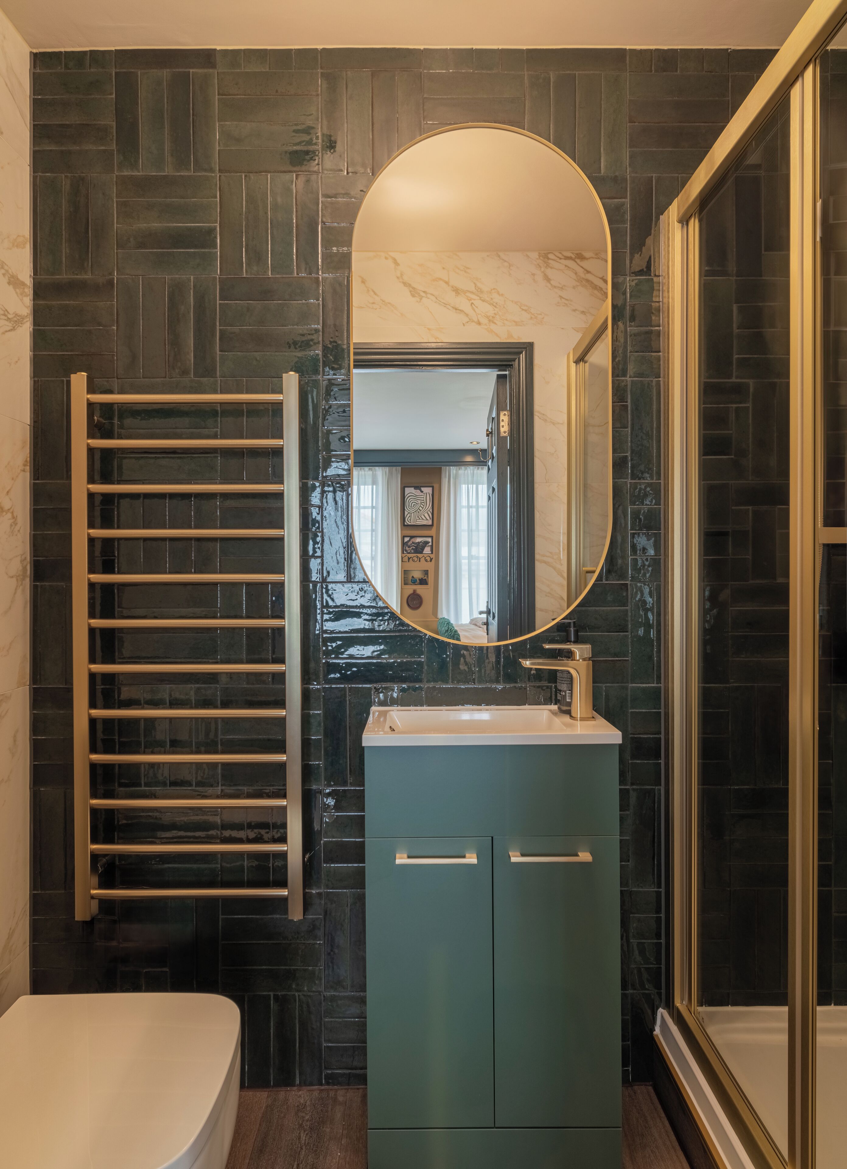 Une salle de bain avec un mur recouvert de carreaux foncés, un lavabo blanc, un meuble vert, un porte-serviettes et un grand miroir.