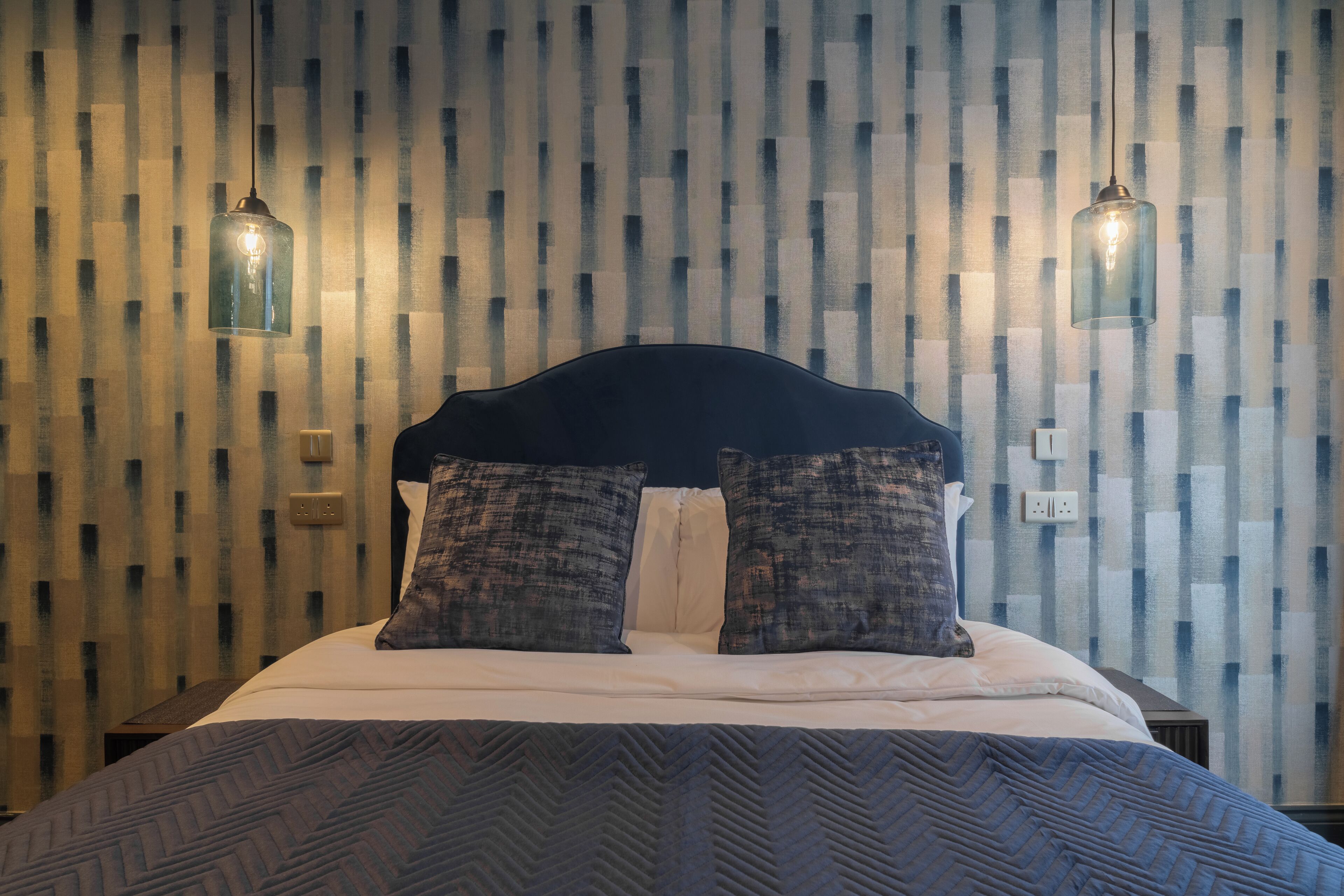 Una cama bien hecha con un cabecero oscuro, dos almohadas estampadas y una colcha estampada. Hay dos lámparas de pared a cada lado de la cama.