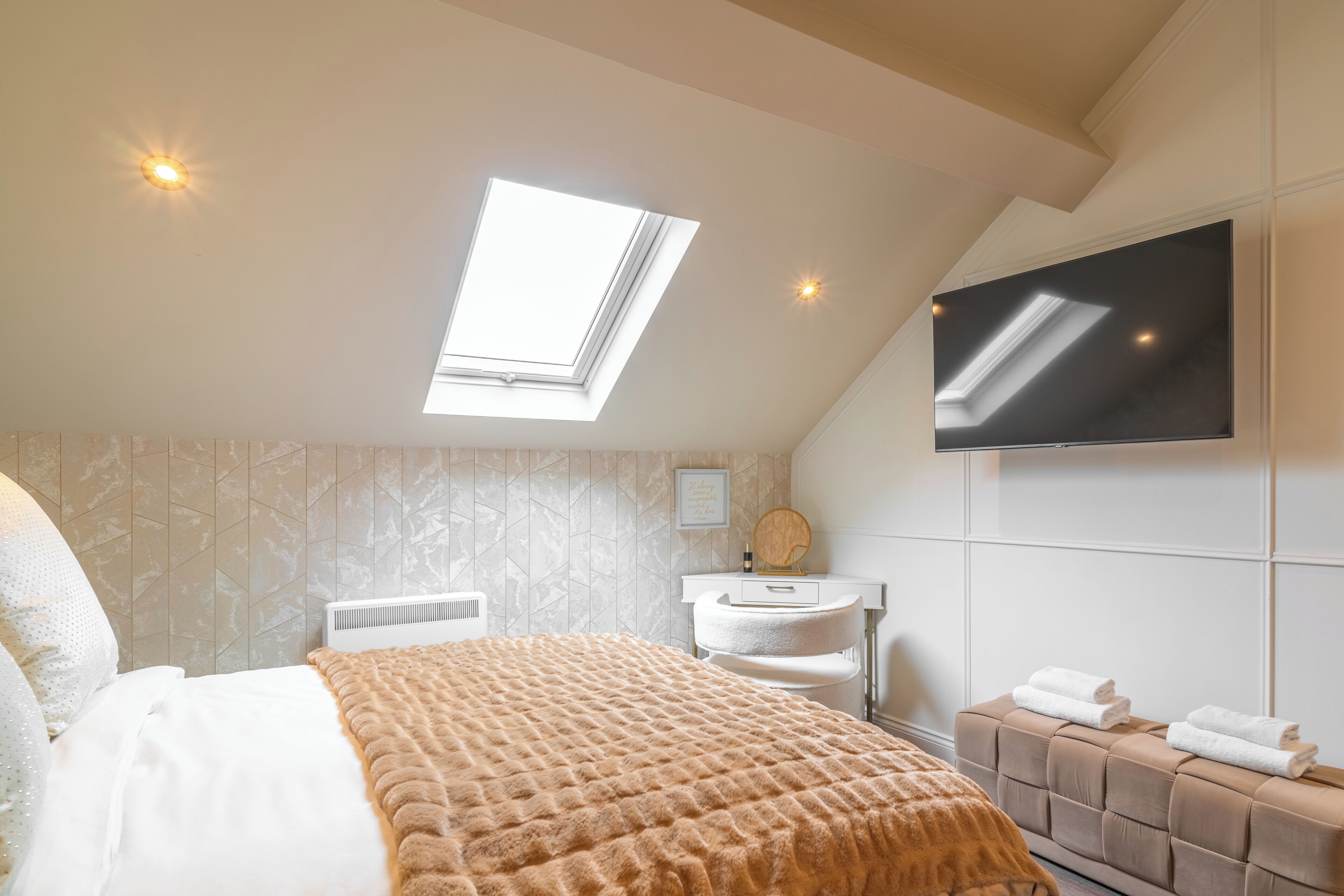 Een slaapkamer met dakraam, een bed met bruine deken, een witte radiator, een kaptafel met spiegel en een aan de muur bevestigde televisie.