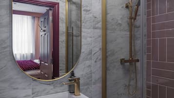A bathroom with a marble wall, a round mirror with a gold frame, a white sink, and a glass-enclosed shower area.
