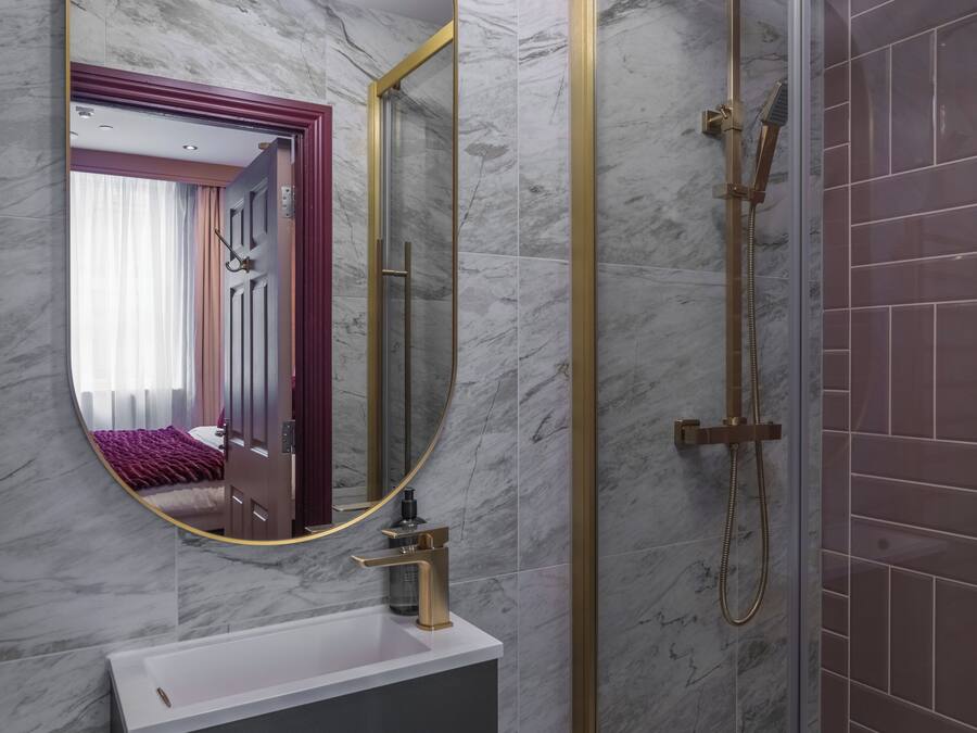 Un baño con pared de mármol, un espejo redondo con marco dorado, un lavabo blanco y una zona de ducha con mampara de cristal.