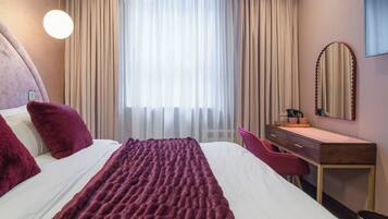 Quarto de hotel com uma cama, uma escrivaninha, uma cadeira, um espelho e uma janela com cortinas.