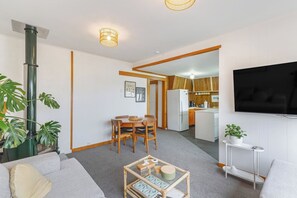 A compact living space with a sofa, a coffee table, a dining area with chairs, a kitchen with a refrigerator, and a flat-screen TV mounted on the wall.