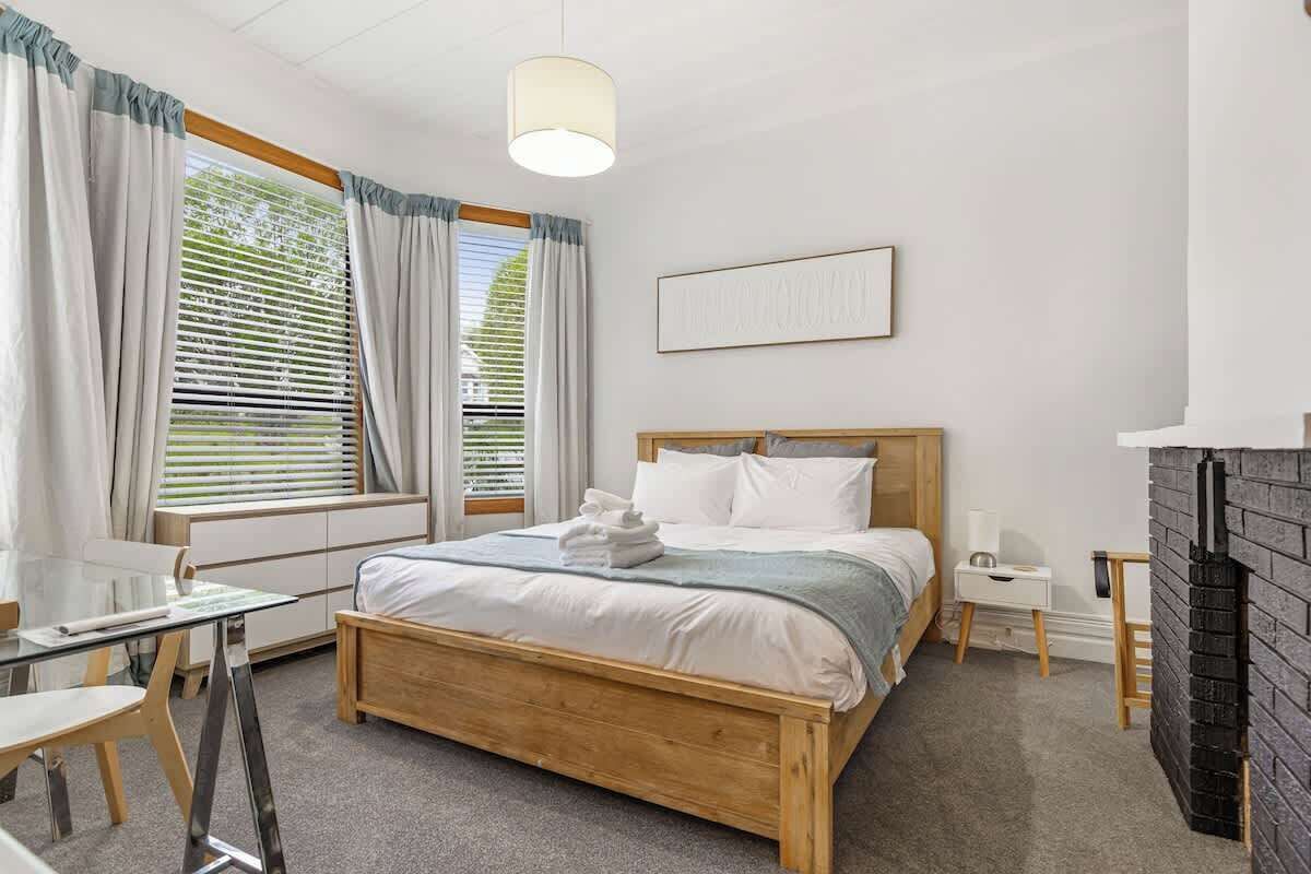 A bedroom with a wooden bed, a glass table, a white bedside table, a fireplace, and a window with curtains.